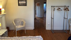 A room with a white wicker chair, a bed with a patterned bedspread, a framed picture on the wall, and a metal drying rack with hangers.