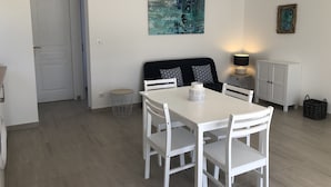 Un coin repas avec une table et des chaises blanches, un canapé noir agrémenté de coussins à motifs, un tableau au mur et une lampe posée sur une table d’appoint.