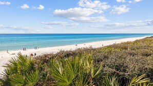 A beach with clear blue water, sandy shore, and green vegetation in the foreground. - Coastal Pet-Friendly Home w/ Brand New 1st Floor Deck | Near Lagoon Pool & Beach (Panama City Beach)