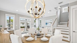 A dining area with a round table, white chairs, a chandelier, and a staircase. - Coastal Pet-Friendly Home w/ Brand New 1st Floor Deck | Near Lagoon Pool & Beach (Panama City Beach)
