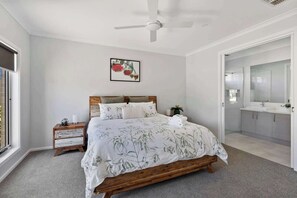 A bedroom with a wooden bed, a nightstand, a bedside table, a ceiling fan, a framed picture on the wall, and a bathroom with a mirror and sink.