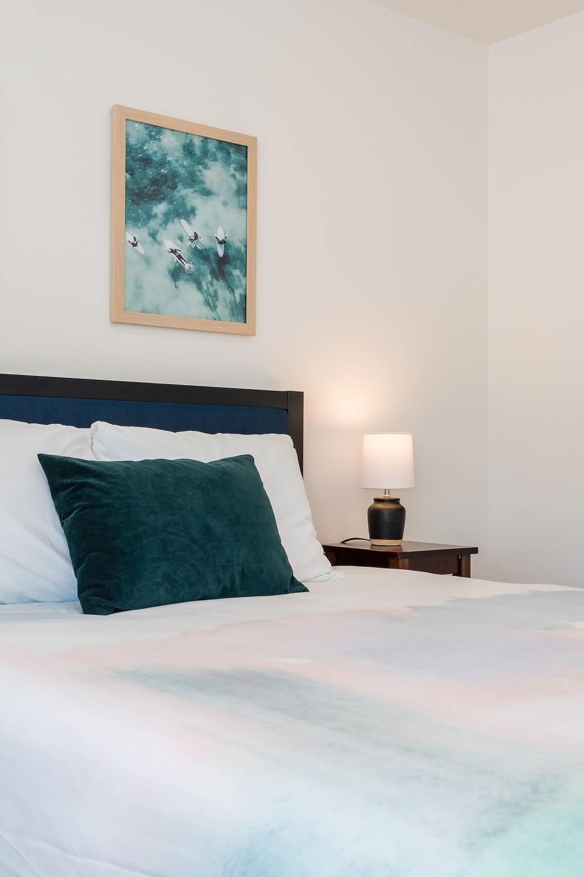 A bed with white and teal bedding, a dark headboard, and a teal pillow. A framed artwork of surfers is on the wall, and a bedside lamp is on the nightstand.