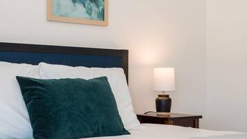 A bed with white and teal bedding, a dark headboard, and a teal pillow. A framed artwork of surfers is on the wall, and a bedside lamp is on the nightstand.