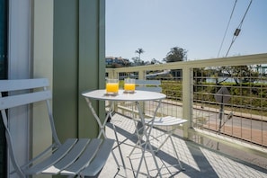 A balcony with a white metal table and chairs, two glasses of orange juice, and a view of a road and greenery.