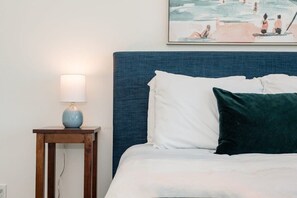 En seng med hvide og mørkegrønne puder, et blåt sengegavl og et natbord med en lampe.