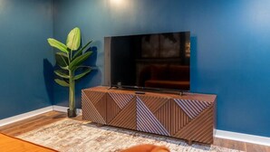 Sala de estar com uma grande TV de tela plana na parede, um armário de madeira com padrões geométricos, uma planta verde grande e um tapete estampado.
