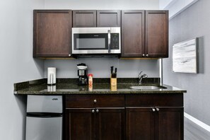 Une cuisine compacte avec des meubles de couleur bois foncé, un four à micro-ondes en acier inoxydable, une machine à café, un évier et un petit réfrigérateur.