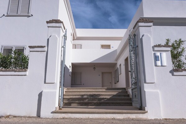 A building with a central staircase leading to a door, flanked by white walls and windows with shutters.