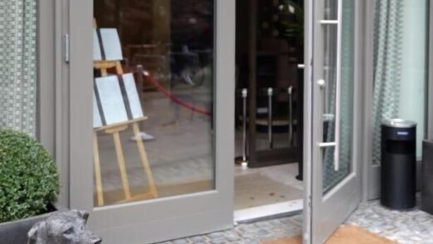 A modern building entrance with a glass door, a small outdoor seating area, and a statue of a dog. - Clounys Hotel & Restaurant (Hamburg)