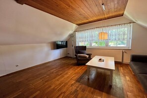 A room with a wooden ceiling, a TV, a chair, a table, and a window with curtains.
