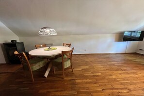 A dining area with a round table, four chairs, a hanging light fixture, a television mounted on the wall, and a herringbone-patterned wooden floor.