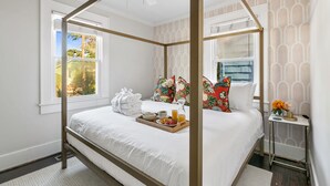 Ein Bett mit Baldachin, ein Tablett mit Frühstücksgebäck, ein Nachttisch mit Lampe und ein Fenster mit Blick auf Bäume.