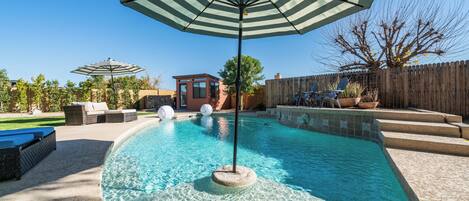 A backyard with a swimming pool, a lounge area with a striped umbrella, and a wooden fence.