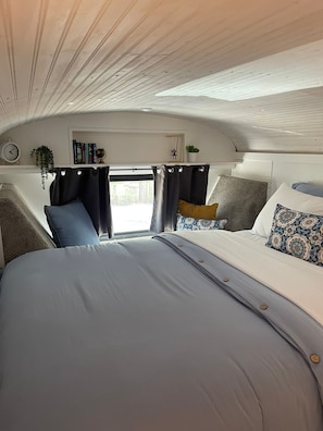 Um interior acolhedor com uma cama, duas mesas de cabeceira, uma estante com livros e uma planta, além de uma janela com cortinas.
