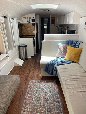 Interior compacto de um veículo recreativo, com cozinha compacta, sofá com almofadas, lata de lixo e tapete estampado.