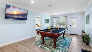 A pool table in a room with a large TV displaying a sunset, a door with a decorative glass panel, and a view of a pool outside.