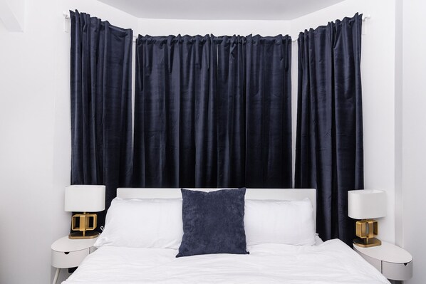 A hotel room with a bed, white bedding, two bedside tables with lamps, and dark curtains.