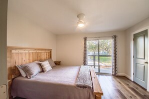 A bedroom with a wooden headboard, a large bed with a blanket, a bench, and a view of the outdoors through a glass door. - River Cliff Retreat Cl 954 7 Bedroom Home by RedAwning (Canyon Lake)