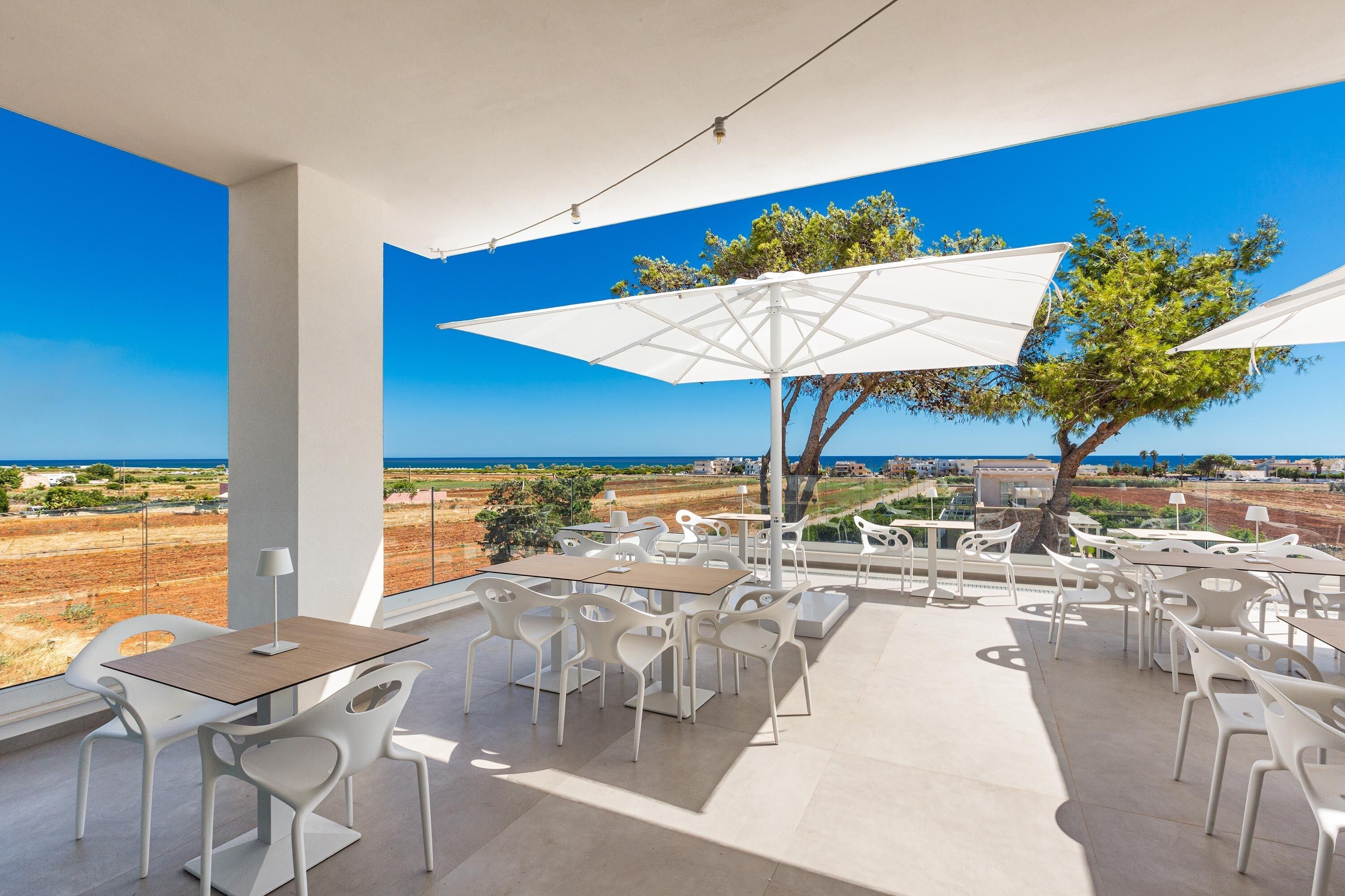 An outdoor patio with white tables and chairs, large umbrellas, and a view of the sea.