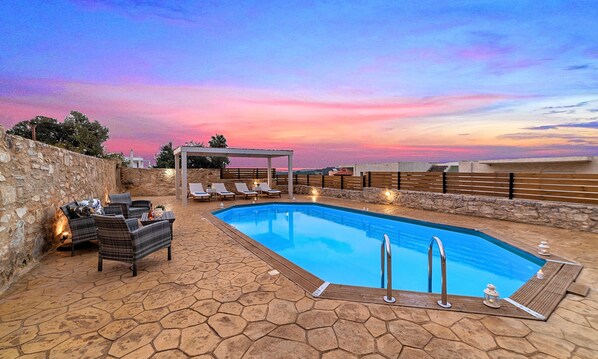 A swimming pool with lounge chairs and a pergola, surrounded by stone walls and a sunset view.