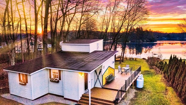 Una cabaña pequeña de un piso con un porche y una pequeña terraza, rodeada de árboles y un lago.