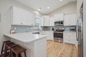 Una cocina con armarios blancos, una isla central, taburetes de madera, microondas, horno y refrigerador.