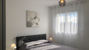 Ein modernes Schlafzimmer mit einem Bett, zwei Nachttischlampen, einer Wandleuchte, einem Fenster mit leichtem Vorhang und einem floralen Kunstwerk an der Wand.