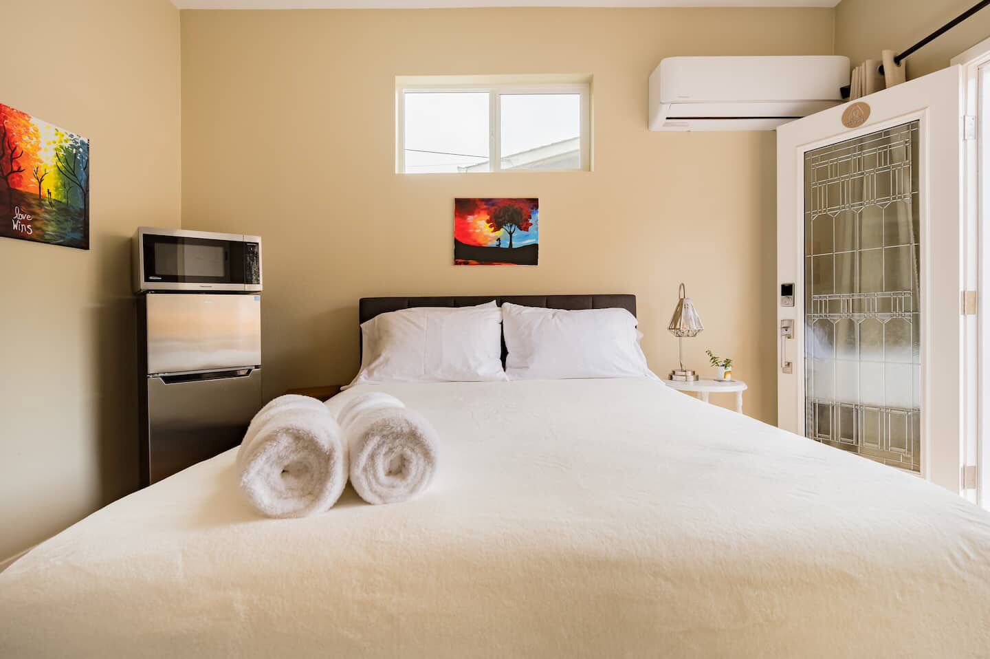 Une chambre d’hôtel avec un lit, deux serviettes enroulées, un four à micro-ondes, une petite table avec une lampe, un tableau au mur et un appareil de climatisation.