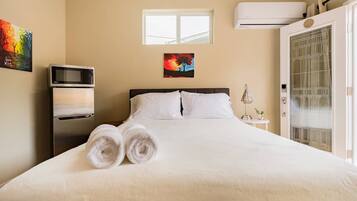 Une chambre d’hôtel avec un lit, deux serviettes enroulées, un four à micro-ondes, une petite table avec une lampe, un tableau au mur et un appareil de climatisation.