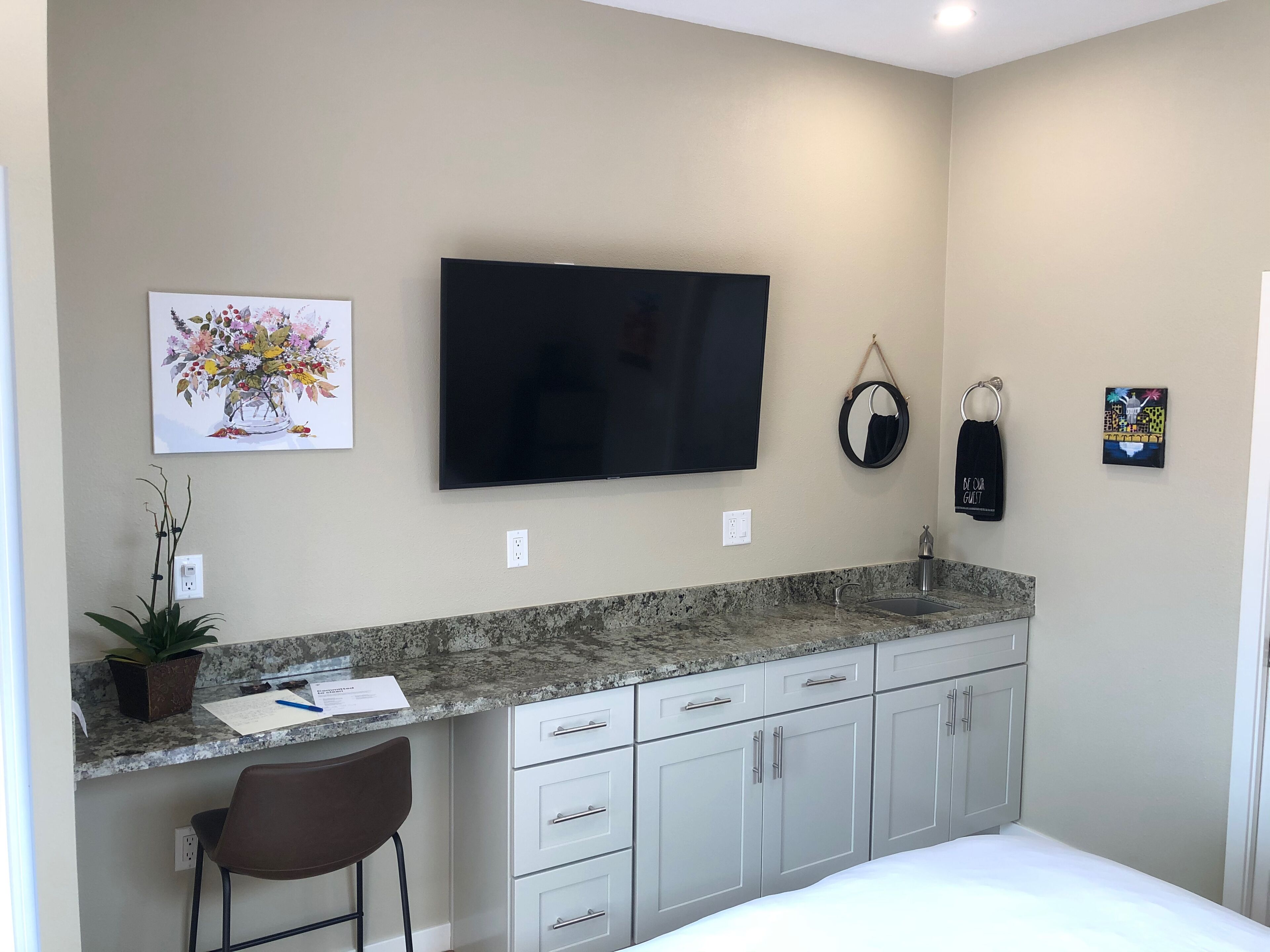 A modern kitchen with a granite countertop, white cabinets, a black TV mounted on the wall, a potted plant, and a small bar stool.