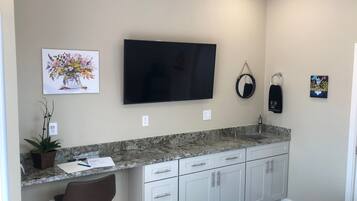 A modern kitchen with a granite countertop, white cabinets, a black TV mounted on the wall, a potted plant, and a small bar stool.
