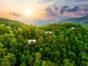 Uma área arborizada com três cabanas, cada uma com um estilo de telhado diferente, cercada por árvores verdes densas e um cenário montanhoso.