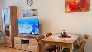 A living room with a wooden dining table, chairs, a flat-screen TV, a clock, and a painting.