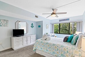 Une chambre à coucher avec un lit, un téléviseur posé sur une commode, un ventilateur de plafond et une fenêtre donnant sur des arbres.