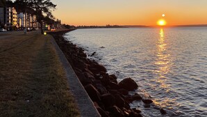 Ein Küsten-Sonnenuntergang mit einem gepflasterten Weg entlang des Wassers, felsigem Ufer und Gebäuden im Hintergrund.