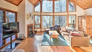 Un vaste salon doté d’un grand téléviseur, de sièges confortables, d’une cheminée et de grandes fenêtres offrant une vue sur la forêt environnante.
