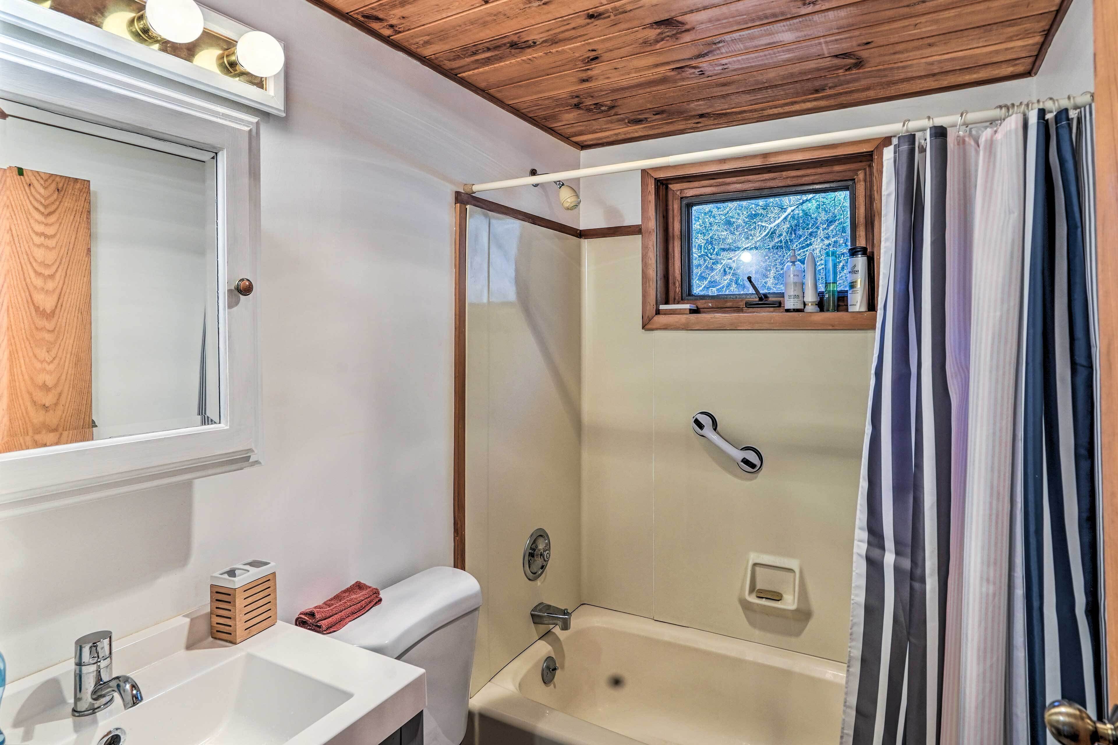 Ein Badezimmer mit Holzboden, einem großen Spiegel, einem Waschbecken, einer Toilette und einer Dusche mit Fenster, sowie einem gestreiften Vorhang.