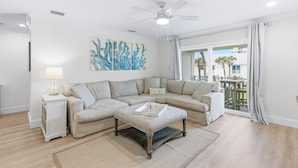 A modern living room with a sectional sofa, a side table with a lamp, a decorative wall piece, and a view through a large window. - Chic, Updated Ocean View Condo on No-Drive Beach (New Smyrna Beach)