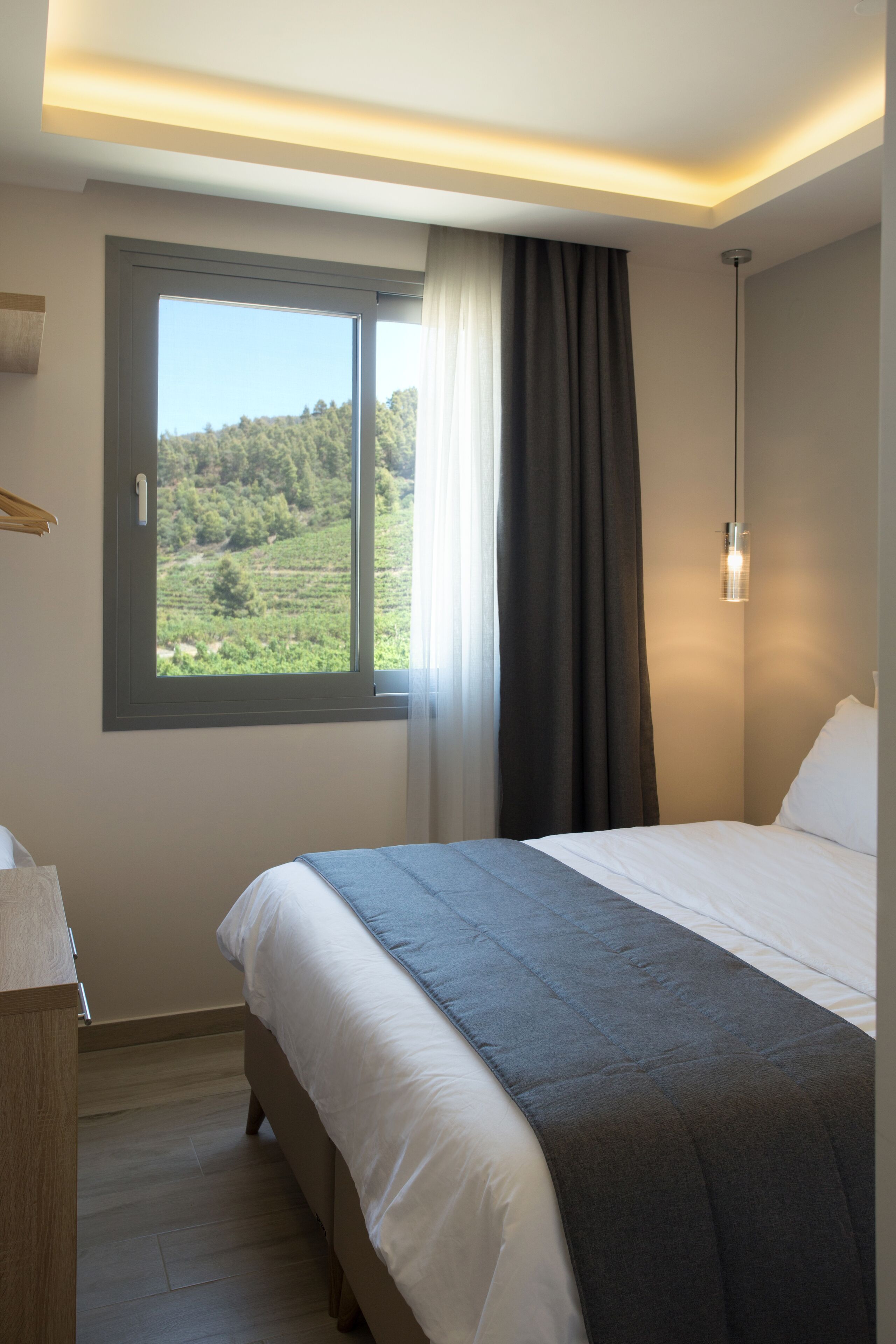 Un dormitorio con cama, ventana con vistas a un paisaje verde, mesita de noche y lámpara colgante.