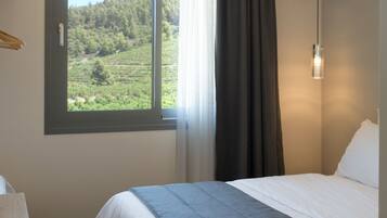 Un dormitorio con cama, ventana con vistas a un paisaje verde, mesita de noche y lámpara colgante.