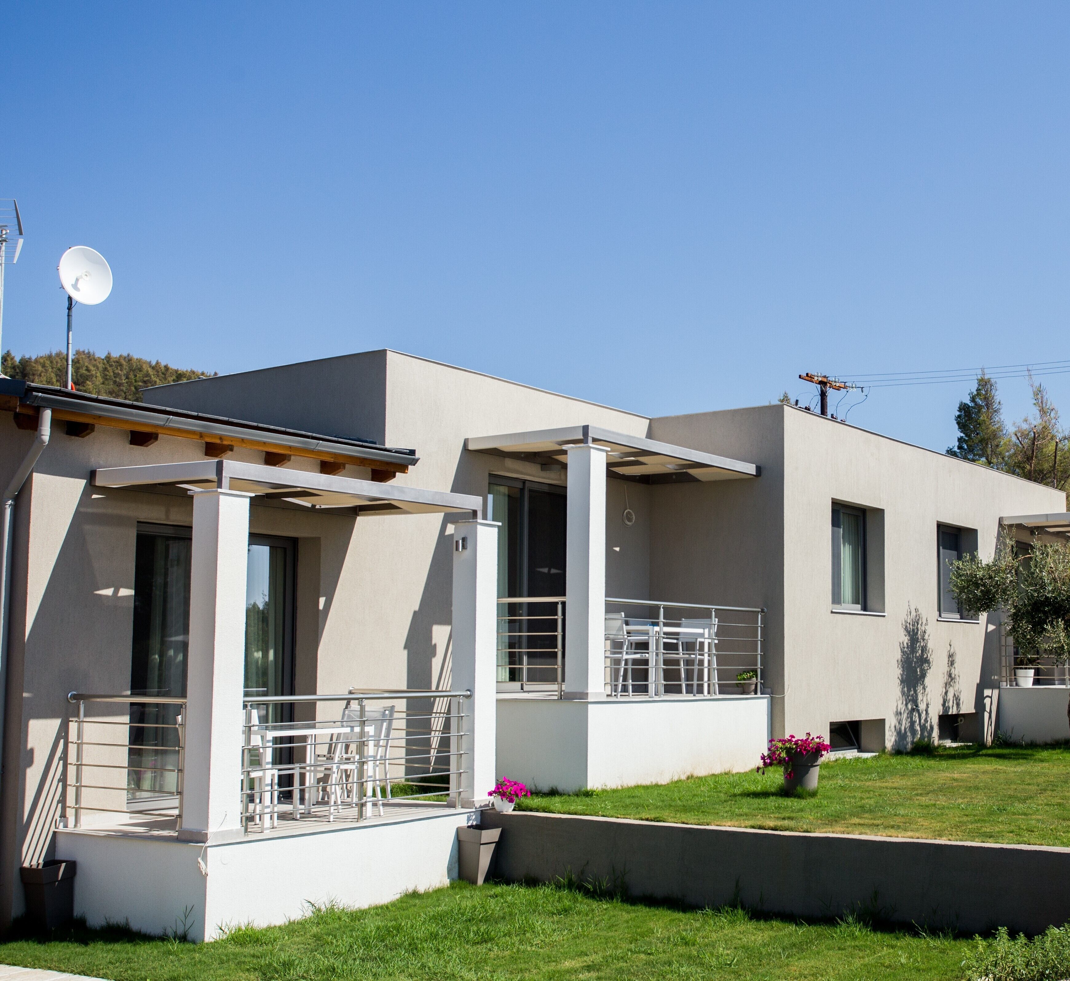 Uma casa moderna de dois andares com um alpendre coberto, antena parabólica e um pequeno jardim florido.