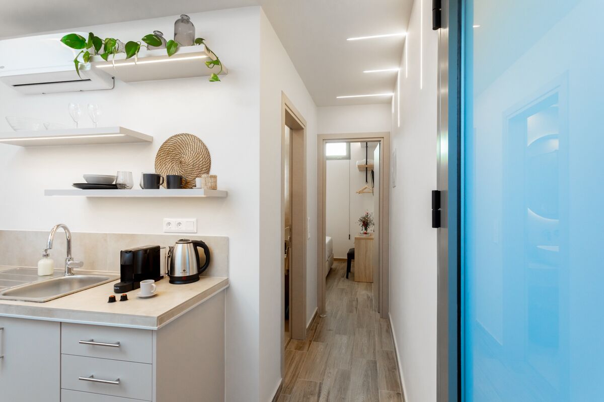Una cocina moderna con armarios blancos, una cafetera y un lavabo. Hay estantes con objetos decorativos y una planta. Se ve una puerta azul a la derecha.