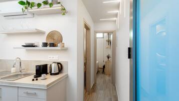 Una cocina moderna con armarios blancos, una cafetera y un lavabo. Hay estantes con objetos decorativos y una planta. Se ve una puerta azul a la derecha.