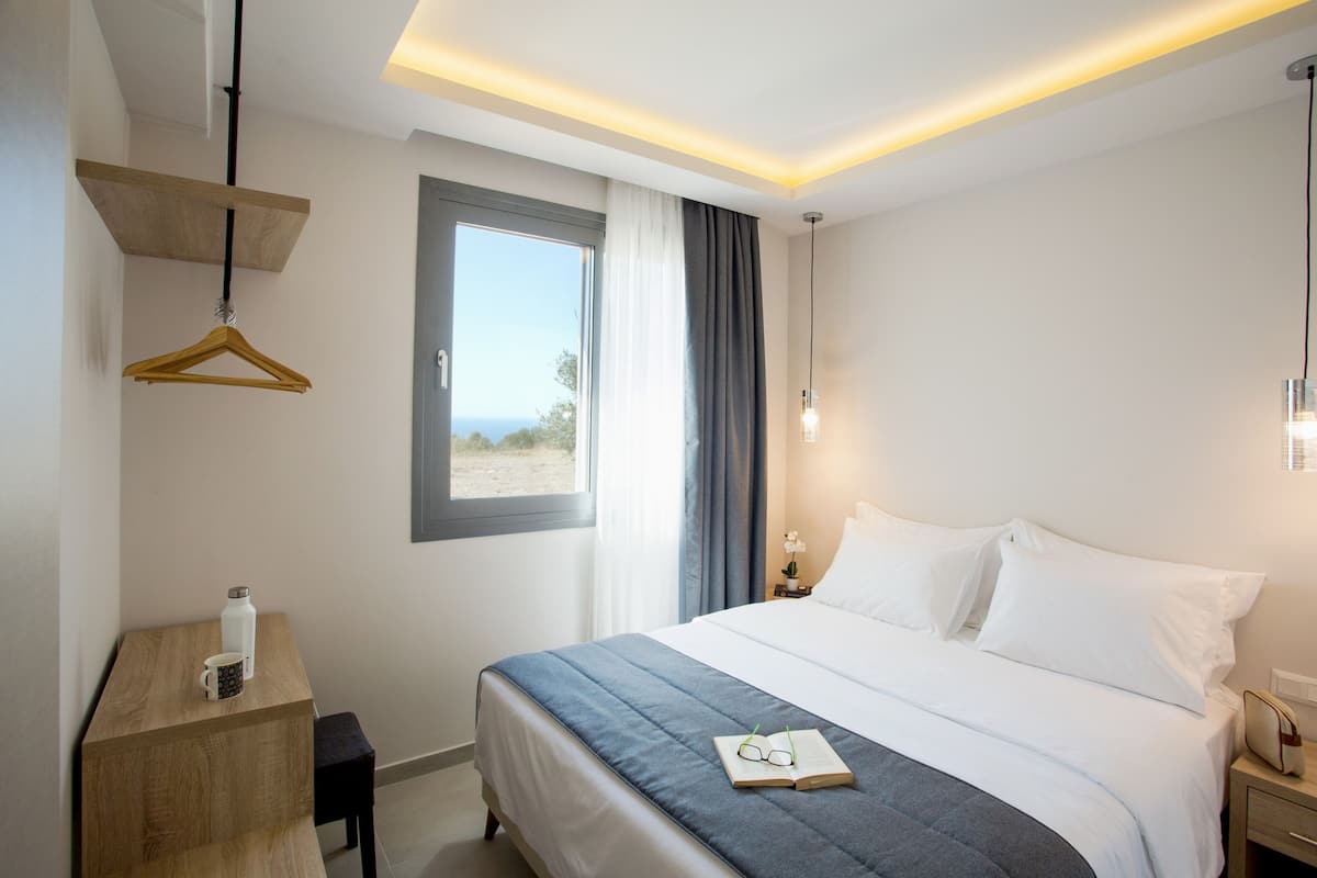A hotel room with a bed, bedside table, a hanging wooden hanger, a window with curtains, and a view of the outdoors.