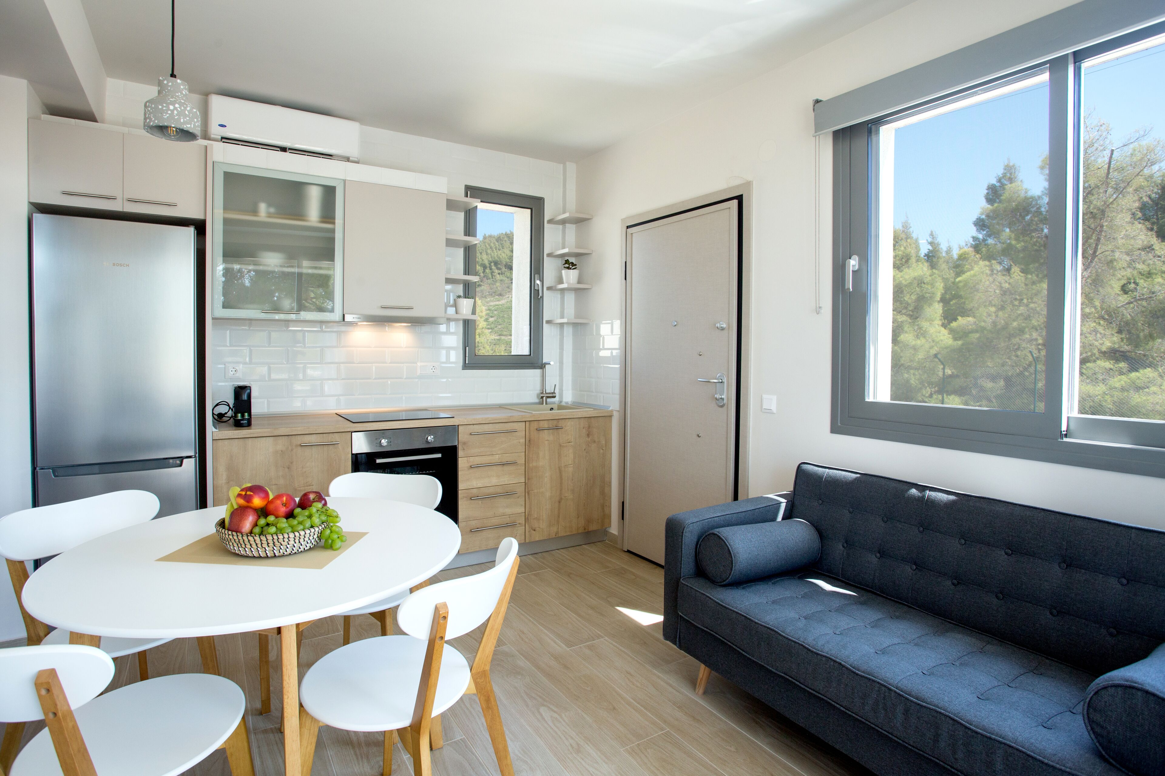 A modern kitchen with white cabinets, a dining table with chairs, a blue sofa, and a large window with a view of trees.