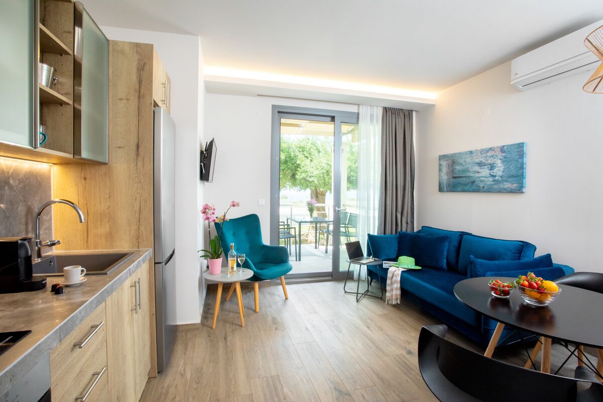 A modern living room with a blue sofa, a round coffee table, and a kitchen area with wooden cabinets and a built-in oven.