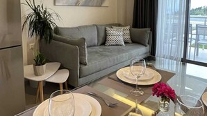 Uma sala de estar moderna com um sofá cinzento, uma mesa de centro com tampo de vidro, e um espaço de jantar com mesa para dois.