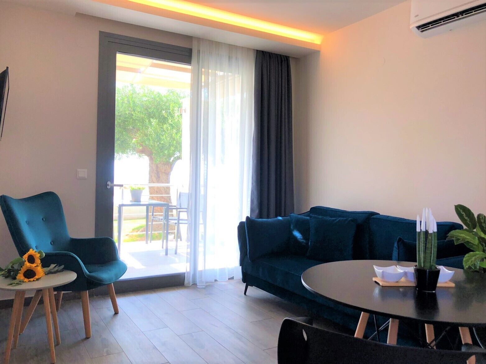 Una sala de estar moderna con un sofá azul, una mesa redonda y una silla. Hay un ventanal corredera que da a un espacio exterior con una mesa y sillas.