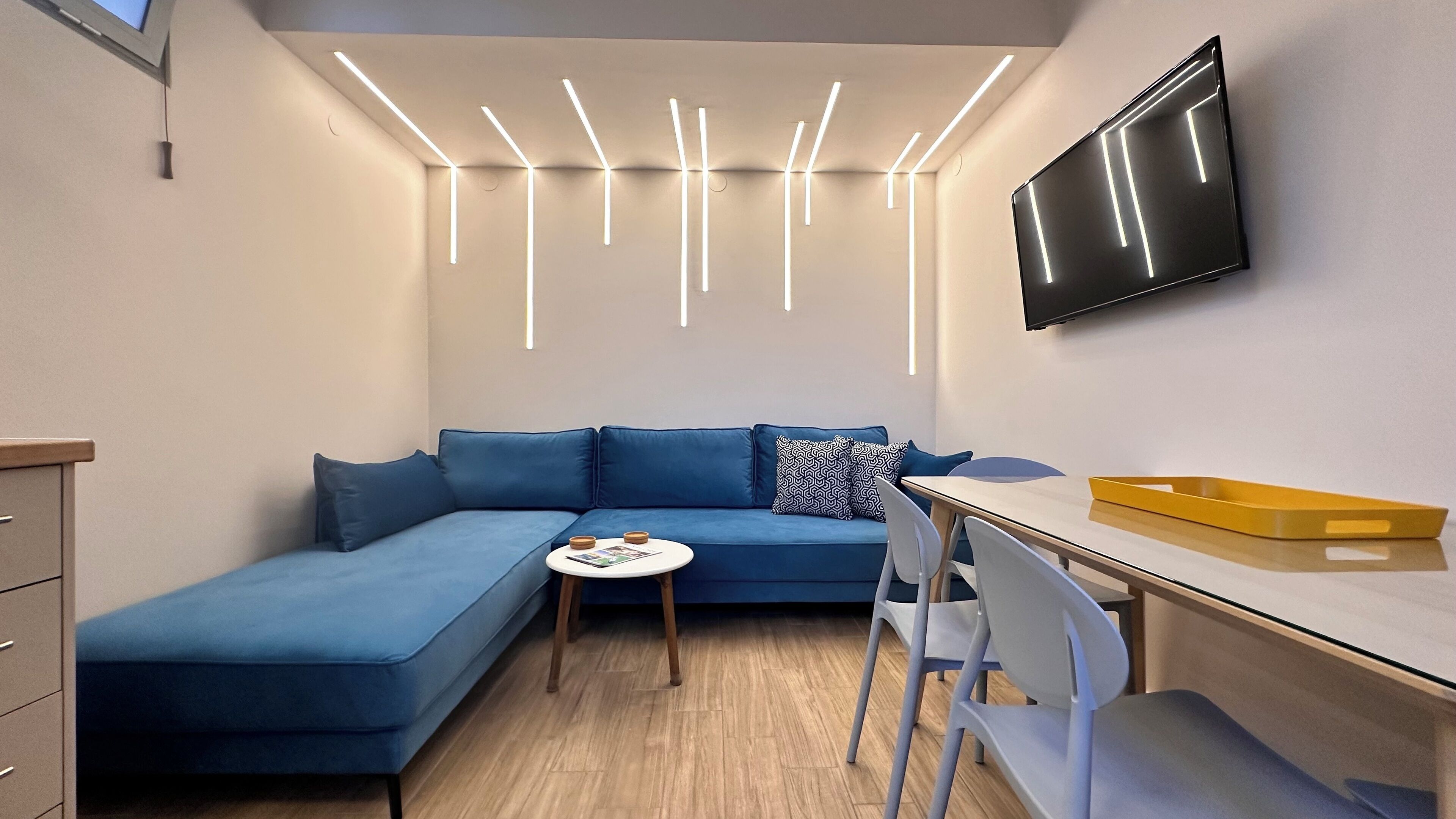 A modern living room with a blue sectional sofa, a small round table, a flat-screen TV mounted on the wall, and a wooden desk with a yellow tray.