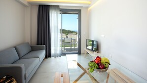 Uma sala de estar moderna com um sofá cinza, uma mesa de centro de vidro com frutas, uma televisão e vista para uma varanda com móveis de exterior.